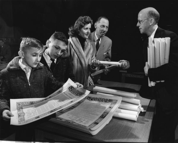 A man is handing a rolled document to a woman. Another man is standing next to her on the right, and two boys are on the left. More rolled documents are resting on a table between them, along with unrolled documents, which the boys are examining. Caption reads: "<b>Copies of the Declaration of Independence</b> were given Tuesday morning to new United States citizens after naturalization ceremonies in the federal court. From left are Vlade Deura, 9, and his brother Stevo, 10, of 1210 S. 89th st., West Allis; Mrs. Maria Jones, 6713 W. Sheridan st.; Dietrich Heinrich Sturm, 4020 N. 23rd st., and Clark Roby, 4364 N. Marlborough dr., Shorewood. Roby, vice-president of the Sertoma Club of Milwaukee, gave the documents on behalf of the club. Federal Judge Robert E. Tehan administered the oath of 88 persons."