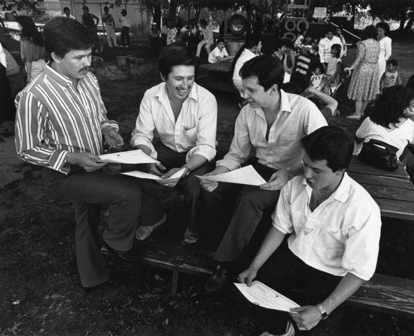 Four men are standing and sitting at a picnic table while holding pieces of paper and talking. Behind them groups of people are relaxing. Caption reads: "COOKOUT CELEBRATION - Fernando Diaz (left) looked over a certificate showing awarded [sic] to students who finished courses in history and English to help them qualify for a US permit card. His brothers, Froglan, Antonio, and Abdon Diaz looked on at a cookout held to celebrate the graduation for a group of students who took the courses La Causa de Esperanza, 410 Arcadian Ave. in Waukesha. Fernando was one of the instructors and his brothers were all in the graduation ceremony." 