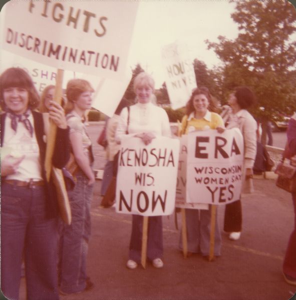 A group of women are standing outdoors holding signs. One sign reads: "Fights Discrimination." Other signs read: "Kenosha Wis. NOW" and "ERA Wisconsin Women Say YES."