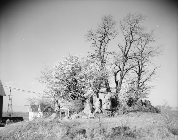 View of several trees growing behind a rocky outcropping. In the background on the left is a farmhouse and farm buildings, and a tower for what may be a windmill. Caption reads: "Pine Bluff (vicinity highway P), Wis. May 13, 1961. Rocks and early spring trees in front of farm house."