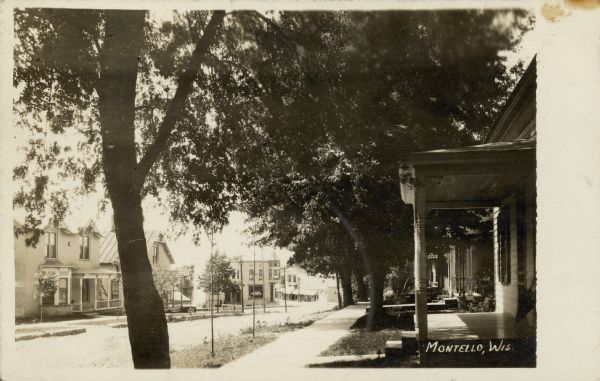 Photographic postcard view looking west down Barstow (now East Montello) Street.  Trees shade the houses at right. The two-story brick home of Frank Dodge is at far left. The Montello State Bank building with its corner turret is at center, with a glimpse of the business district beyond. Caption reads: "Montello, Wis."