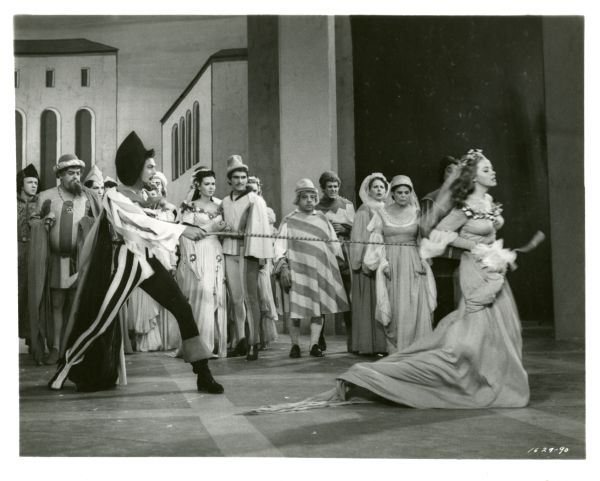 A scene still from the 1953 film "Kiss Me Kate" starring Howard Keel and Kathryn Grayson. In the scene, Keel, as Petruchio, has snared Kate, Grayson, around the waist with a whip as she tries to leave. A crowd of costumed actors are looking on.