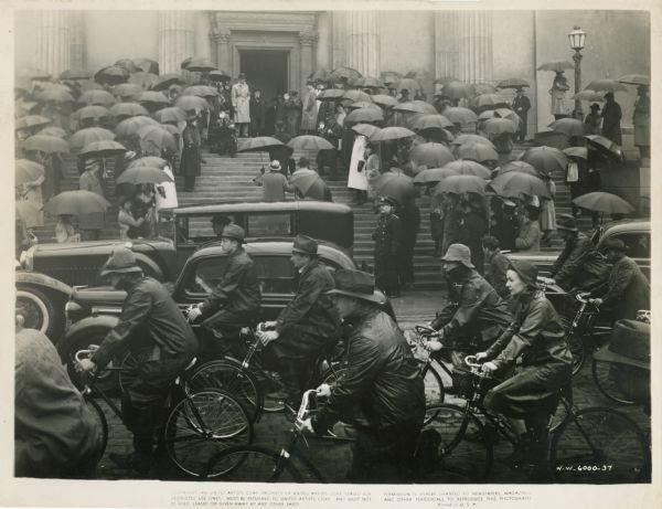 Scene still from the 1940 film "Foreign Correspondent." Cars and people on bicycles pass a large crowd of people under umbrellas standing on the steps of a building in the rain. Several men are walking up the steps towards an open door. Several photographers and a few men wait at the top of the steps.