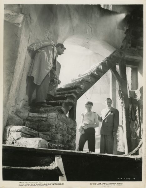 Scene still from the 1940 film "Foreign Correspondent." Mr. Krug (Eduardo Ciannelli) stands with his hand on the shoulder of Tramp (Martin Kosleck) underneath a staircase inside a windmill. John Jones (Joel McCrea) stands on the steps of the stairs to the left of the two and tries to stay out of sight.