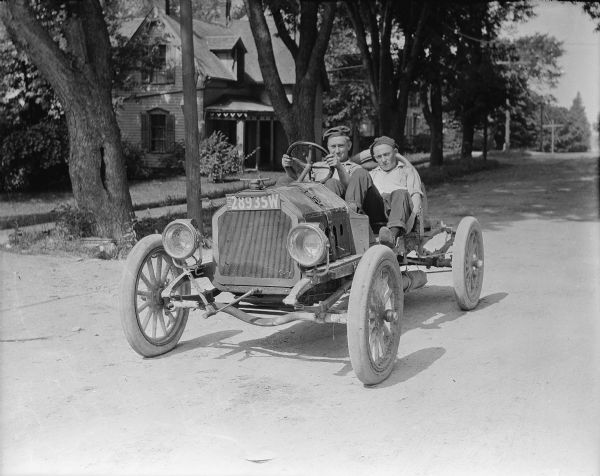 Two youths are sitting in what appears to be a homemade racecar on a residential street. There is a 1913 license plate on the front of the car. A house is in the background.