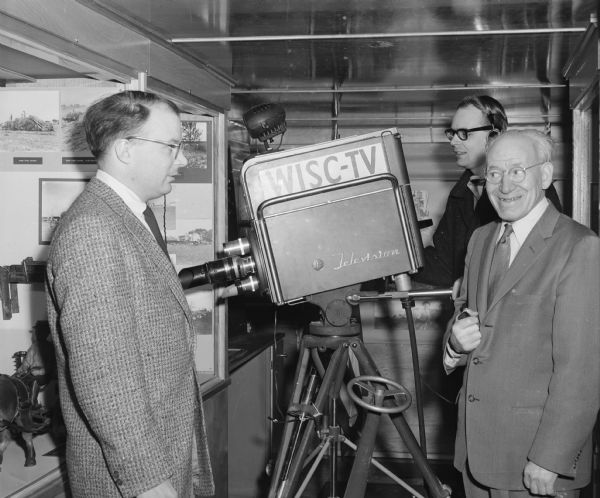 Two men wearing suits are standing in the foreground, with the man on the right holding a pipe in his hand. Behind them is a man standing and operating a WISC-TV television camera which is aimed towards a display case. Caption reads: "Walter Dunn and others inside Historymobile; television broadcast."