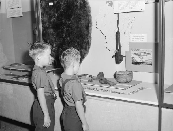 Two children are standing and looking at an exhibit that features tools, a map, and some text. Caption reads: "Visitors in the pioneer gallery of the State Historical Society of Wisconsin, 1957."