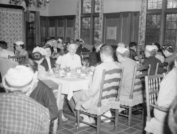 A group of well-dressed men and women are sitting at tables in a well-decorated room. Caption reads: "Kohler, Wis. June 6, 1957. Members of the Women's Auxiliary of the State Historical Society of Wisconsin on their annual pilgrimage, at lunch in the American Club at Kohler Village."