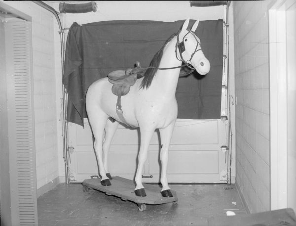 A statue of a horse on a cart in front of a garage door. Caption reads: "Wooden horse presented to the State Historical Society of Wisconsin by the State Medical Society for display at Stonefield Farm and Crafts Museum, 1957."