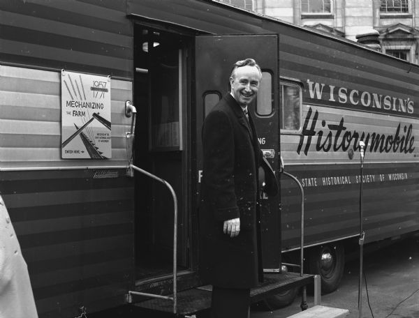 A smiling man is standing on the steps in the open doorway of a large vehicle painted with a sign that reads: "Wisconsin's Historymobile, State Historical Society of Wisconsin." A sign for a program entitled "Mechanizing the Farm" is posted next to the door.