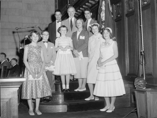 Group portrait of people posing together on steps leading up to a podium. There are three men at the top, and six young people in front. Flags are behind them, and people are sitting people on the left. Caption (from negative envelope) reads: "Rennabohm [sic] Winners at first Annual State Awards Day, May 25, 1957."