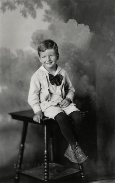 Studio portrait of Gaylord Nelson as a young boy. He is sitting on a table in front of a painted backdrop.