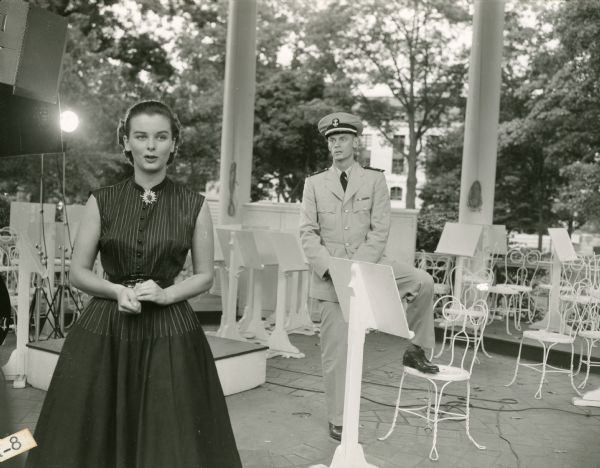 Actress Audrey Dalton and actor Carlton Carpenter film a scene for the television show "Men of Annapolis" on the bandstand at the U.S. Naval Academy.  Carpenter has a foot resting on a chair and is wearing a Navy uniform.