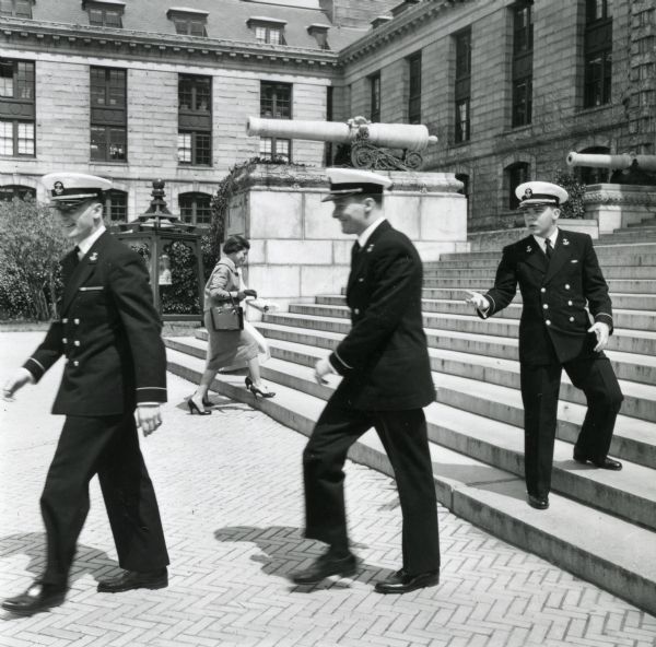 Three actors from the television show "Men of Annapolis" are seen outside of Bancroft Hall at the U.S. Naval Academy. Two men are walking away from the building while the third stands on the bottom steps and looks back at the other two. A woman starting to walk up the steps is seen in the background.