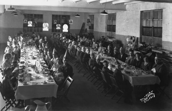 Group portrait of Gardner Baking Co. families wearing paper party hats sitting at two long banquet tables. There are Purity Bread signs over the tables which are hanging from lines strung between the ceiling lamps.