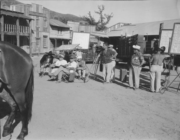 Photo taken on the set of the TV show The Cisco Kid while filming at the Paramount Ranch. A man on a horse is in the foreground on the right and a wagon is on the left. Building facades and light reflectors are seen in the background.