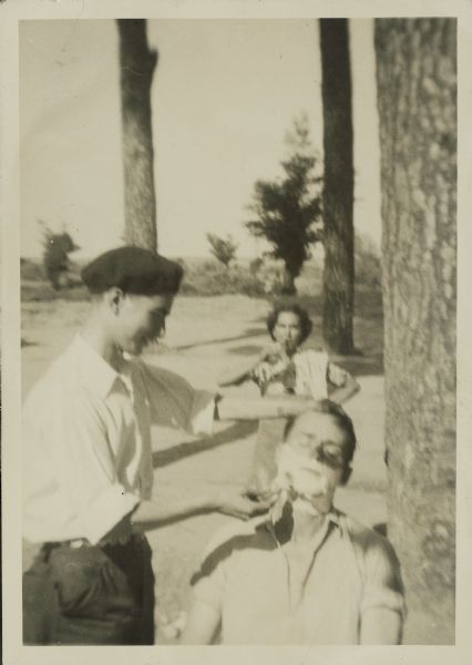 Fred Palmer getting shaved by a man wearing a hat outdoors. Another person is standing in the background.