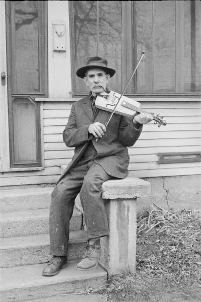 A man is sitting on a front stoop of a building playing a violin. He is wearing a hat and has a pipe in his mouth.
