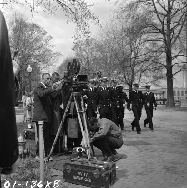 A film crew for the Ziv television show "Men of Annapolis" stand behind and below a camera. A group of naval cadets in uniform can be seen marching in formation behind them. An equipment case marked "ZIV TV BATTERY CASE" is on the ground in front of the camera.