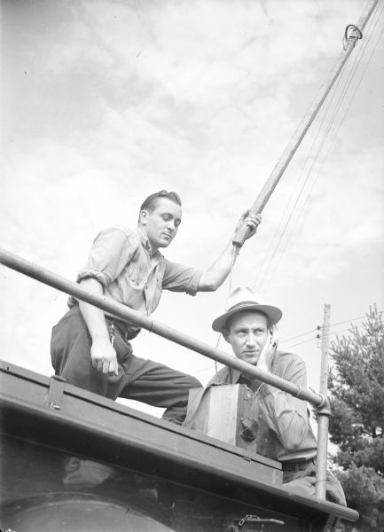 Two men outdoors with an emergency telephone. The men appear to be on top of a truck or other type of vehicle. Behind them are power lines and trees.