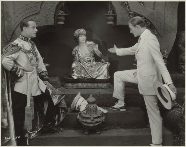 Still from the 1921 film "Fool's Paradise". Arthur Phelps (Conrad Nagel) gestures toward Samaran (Julia Faye) who is seated cross-legged on a small platform in an alcove. Talaat-Ni (John Davidson), a  prince of Siam, stands opposite and looks intently at Phelps — Samaran is his chief wife. The prince wears a uniform with a long cape and decorations.