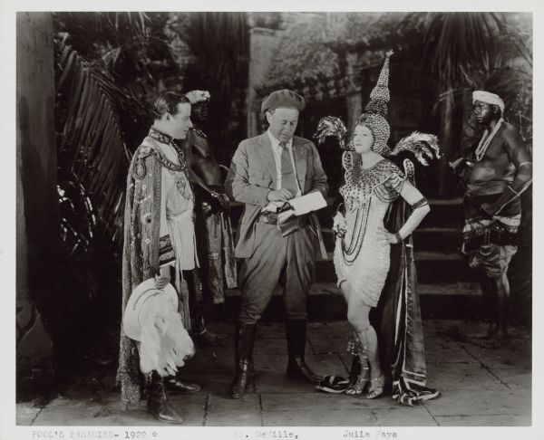 Director Cecil B. DeMille confers with actors John Davidson and Julia Faye on the set of the 1922 film "Fool's Paradise". Davidson is dressed in a uniform and heavily adorned cape. Faye wears a Southeast Asian inspired costume including a conical shaped headdress. Both Davidson and Faye are playing Southeast Asian characters: Prince Talaat-Ni and one of his wives, Samaran. Two African American extras stand in the background.