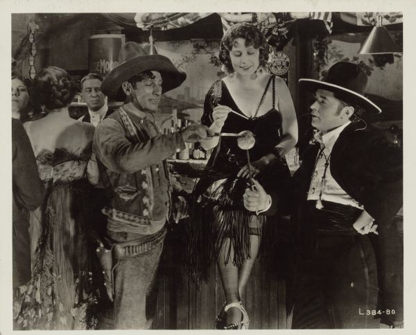 Poll Patchouli (Dorothy Dalton) sits on a bar in a scene from the 1921 film "Fool's Paradise". John Rodriguez (Theodore Kosloff) and another man (George C. Fields) stand on either side of her. Kosloff holds something on a stick and Fields uses a knife to slice the top off.