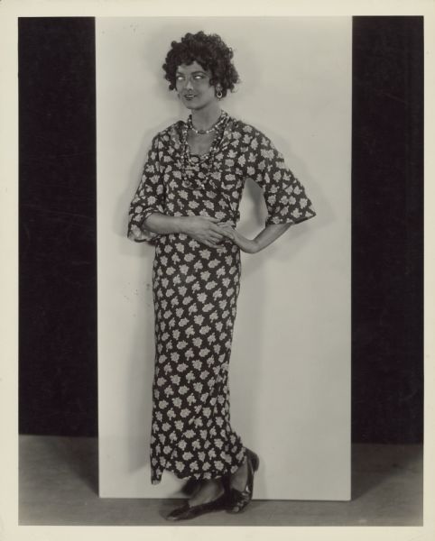 Myrna Loy, who played the Sengalese spy Fifi in the 1927 film "Ham and Eggs at the Front", poses for a photograph. Loy is wearing a black curly wig and blackface makeup wherever her skin is not covered by clothing. She wears a floral print dress with beaded necklace and earrings. 