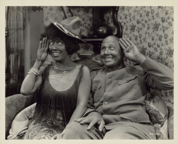 Cally Brown (Louise Fazenda) and Ham (Tom Wilson) sit next to each other on a small sofa in a scene from the 1927 film "Ham and Eggs at the Front". They are both smiling and both have their outside hands to their foreheads in a salute. Ham is dressed in a uniform and Cally wears his hat. She wears a curly wig and both wear blackface makeup.