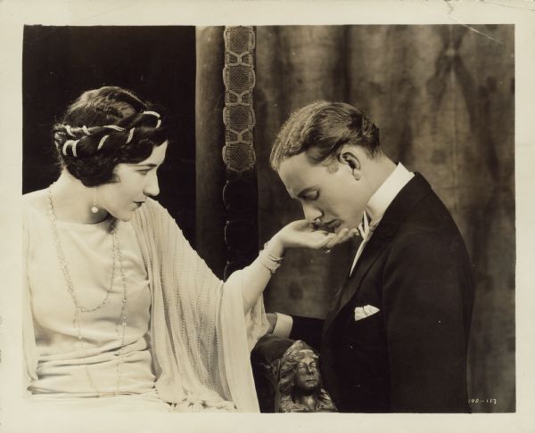 The Queen (Aileen Pringle) looks at Paul Verdayne (Conrad Nagel) as he kneels next to her chair in a scene from the 1924 film "Three Weeks". His head is bent and her hand is under his chin as if she is about to lift his head up.