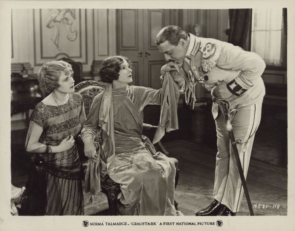 Prince Gabriel (Marc McDermott) kisses the hand of Princess Yetive (Norma Talmadge) as Dagmar (Wanda Hawley) kneels beside her and looks on in a scene from the 1925 film "Graustark". The prince wears a dress military uniform and holds onto a sword as he bends over to kiss Yetive's hand.