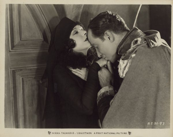 Grenfall Lorry (Eugene O'Brien) kisses the hand of Princess Yetive (Norma Talmadge) in a scene from the 1925 film "Graustark". Her mouth brushes his forehead. The princess is dressed in black while Lorry wears a heavy cape or coat.