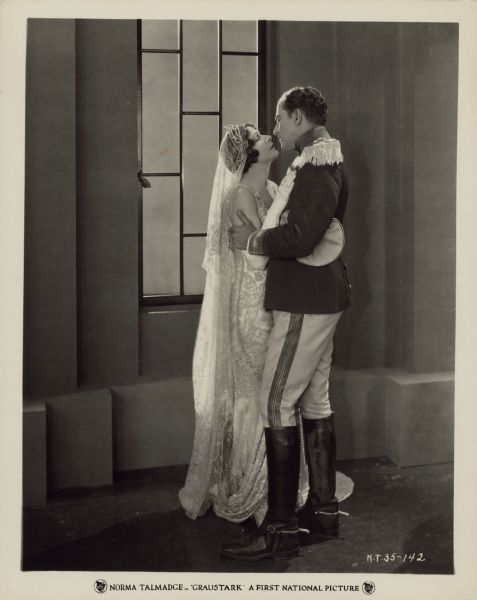Grenfall Lorry (Eugene O'Brien) and Princess Yetive (Norma Talmadge) embrace in a scene from the 1925 film "Graustark". Yetive wears a wedding dress and veil while Lorry wears a uniform with epaulets and riding boots.