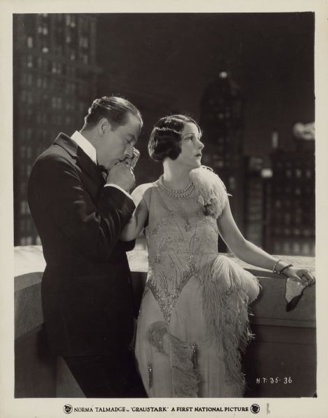 Grenfall Lorry (Eugene O'Brien) kisses and nuzzles the hand of Princess Yetive (Norma Talmadge) in a scene from the 1925 film "Graustark". The two are dressed in formal wear and stand on a balcony of a building. Yetive looks away from Lorry.