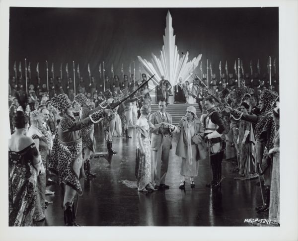 Director Jack Conway and screenwriter Elinor Glyn talk to Eleanor Boardman and Conrad Nagel on the set of the 1925 film "The Only Thing". The four stand underneath the raised swords of soldiers standing on either side of them along with many extras. The soldiers wear leopard print hats and wraps.