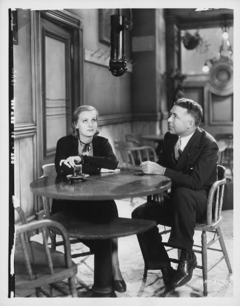Greta Garbo and director Clarence Brown sit at a table on the set of the 1930 film "Anna Christie". They both are looking up at a microphone.