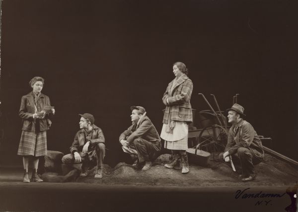 Five cast members of the play "The Eve of St. Mark", including Aline MacMahon and Scott McKay, are seen on stage.  The three men in the cast are dressed in work clothes and all wear hats.  The two women wear skirts, jackets and boots.  They stand on a "mound" of dirt with a wagon behind them; otherwise the stage is bare.