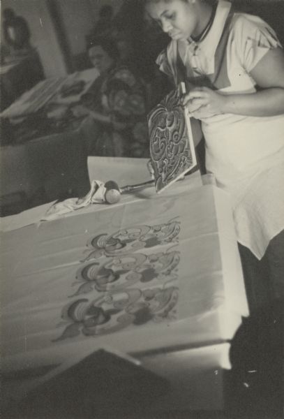 Caption reads: "The material to be printed is stretched over a padded table". Another woman is working at a table in the background.