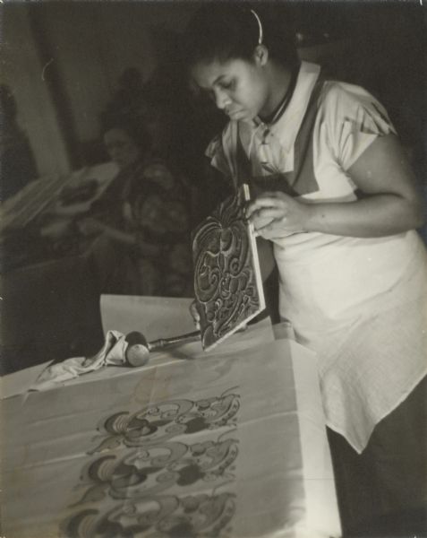 A woman is printing material stretched over a padded table. Another woman is working at a table in the background.