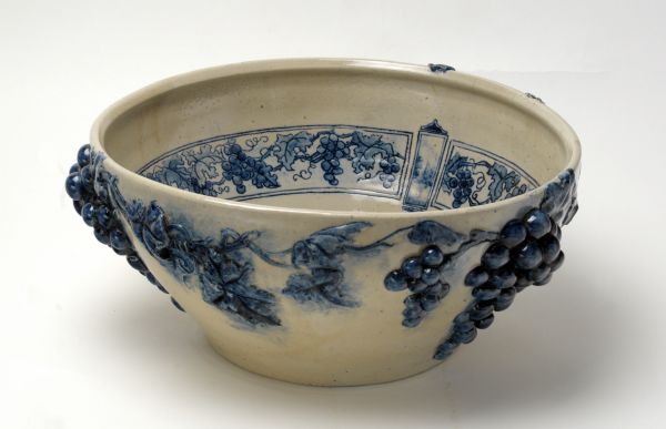 Stoneware bowl with a clear glaze on gray clay. Exterior has raised grape clusters in blue and painted blue underglaze. Interior surface has hand-painted blue grape design motifs. Underside has an incised maker's mark, "SF".