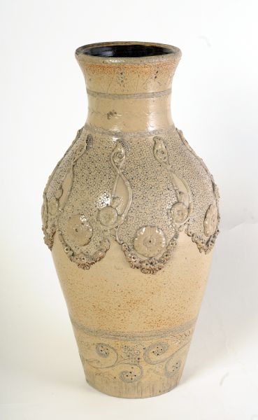 Stoneware vase is ovoid-shaped with a flared rim. It is gray with a salt-glaze. The dark brown on the lower body and the top of the neck may be caused by applying a light wash of iron during the firing to achieve the colored effect. The bottom of the piece is glazed, while the top is not, meaning the piece was likely fired upside-down. The interior of the piece is glazed with a dark brown slip. The rim has a band of incised hatching. The neck has incised leaves and vines, a band of hatching, two bands of intersecting circles, and another band of hatching. The upper body has small stamped quatrefoils and applied flowers, festoons, and floral s-shaped motifs. The lower body has a band of hatching, incised foliate scrolls, and long incised lines near the base. A maker's mark, "SF", and date, "1893", are incised on the underside.
