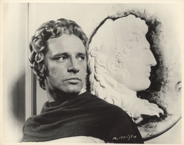Richard Burton poses, in costume, next to a plaque of Alexander the Great.  Burton wears a curly blonde wig for the title role of the 1956 film "Alexander the Great".