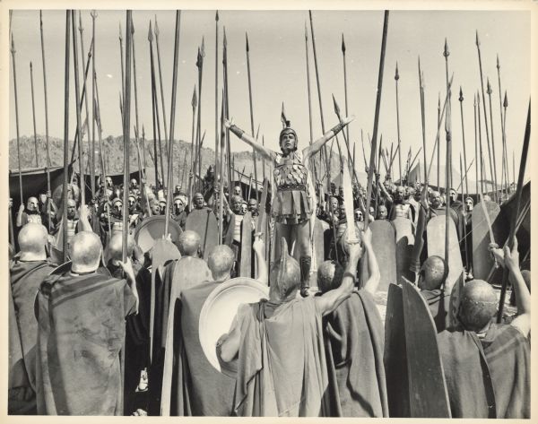 Richard Burton, as Alexander the Great, stands with his arms upraised surrounded by soldiers with shields and very long lances in a scene from the 1956 film "Alexander the Great".
