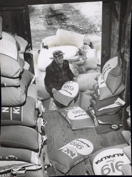 A man lifting a bag either into or out of a shed holding several identical bags. Caption reads: "<b>Each spring Donald Howard,</b> Sussex, makes a trip to the Sussex Mills for his seed supply. Here he loaded alfalfa seed. It will provide next year's hay when planted with this year's oats."