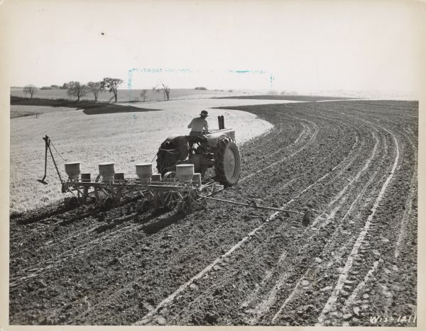 Three-quarter rear view from right of a man driving a tractor with a planter attached. Caption reads: "Wis-1211
Green County, Wis. Gene Rechsteiner, owner, Monticello, Wisconsin

Planting corn in a contour strip with 4-row planter and John Deere big model Tractor. Rechsteiner says large equipment gives no trouble in contour work like this.

SCS Photo by W.H. Lathrop
May 20, 1948"