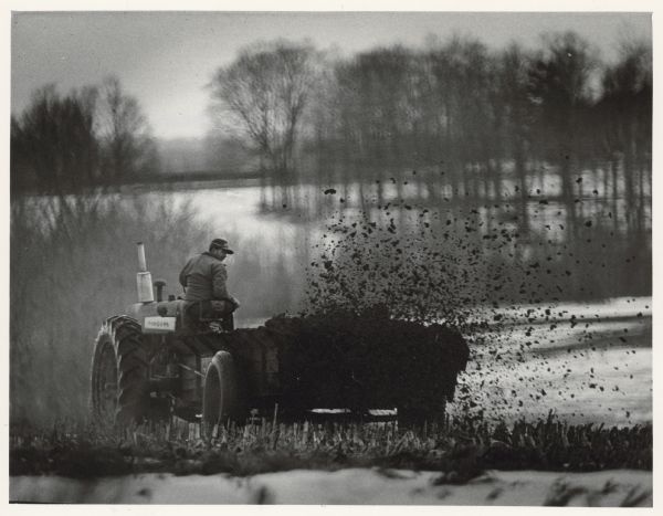 Rear view of a man driving a tractor, which is pulling a manure spreader. Caption reads: "GETTING READY FOR SPRING - A farmer took advantage of a nice winter day to get out into the fields near Athens in Marathon County and spread manure on his land to prepare the ground for spring planting."