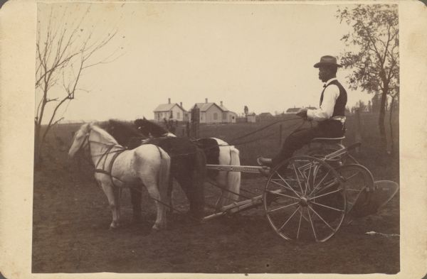 A man is sitting on a plow driven by a team of horses. Buildings are in the distance. Caption identifies the plow as a J. Thompson & Son Company sulky plow, made in Beloit.