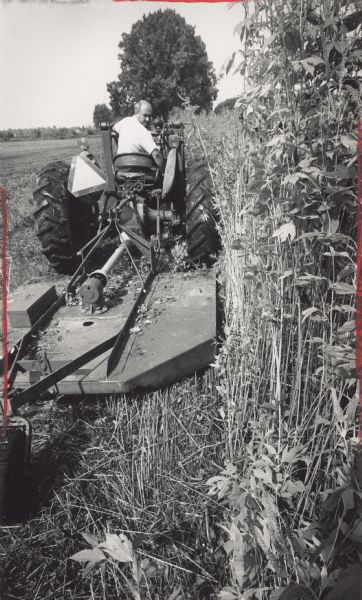 A man driving a tractor that is dragging a mower. Caption reads: "Leonhard Weiss Jr., an Oak Creek weed commissioner, cut 8-foot-tall weeds on Milwaukee County property behind his farm".