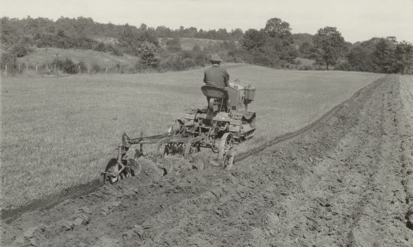 Rear view of a man driving a tractor. An attached plow is plowing the field behind him. Caption reads: "Plowing with Model 'W' 12-20 H.P. [horsepower] Cletrac with three 12" bottom mouldboard plow. Plowing done at 3 1/2 miles per hour. Note the pulverization of soil and complete burial of trash obtained at this speed." 