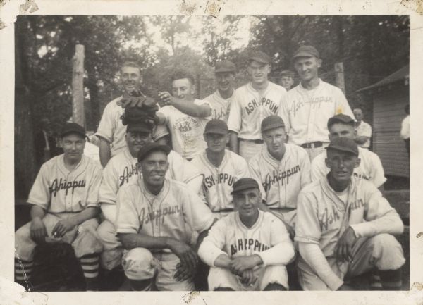 Outdoor group portrait of men wearing baseball uniforms that read: "Ashippun". Back row, left to right: Roy Wittnebel, Floyd Clemens, Norm Nelson, Vic Liesner and Hilbert Linders. Middle row, left to right: unknown, Don Chapman, Don Christianson, Milton Mass, unknown. Front row, left to right: Elmo Wendorf, Ruby Haidman and Ed Wendorf,